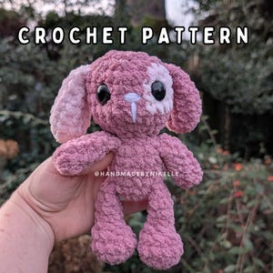 May include: A handmade pink crochet bunny with black eyes and white accents. The bunny has long ears and is made with a textured yarn. The text "CROCHET PATTERN" is at the top of the image, and "@HANDMADEBYNIKELLE" is at the bottom.