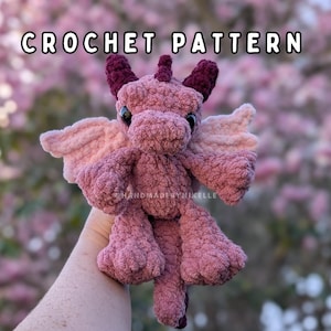 Könnte beinhalten: Ein handgefertigter, gehäkelter Plüschdrache in Rosa- und Burgundertönen. Der Drache hat Flügel, Hörner und einen Schwanz. Der Text "CROCHET PATTERN" wird oben im Bild angezeigt. Der Drache wird in einer Hand gehalten.