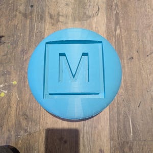 Op de afbeelding: Een rond, lichtblauw object met een vierkante uitsparing in het midden. De letter "M" is in de uitsparing gegraveerd.