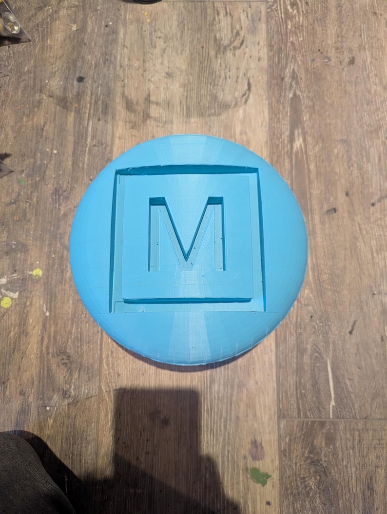 May include: A light blue round object with a square cutout in the center. The letter "M" is carved into the cutout.