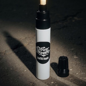 May include: A white paint marker with a black cap and a black circular label featuring the white text "ON THE STREET" and "Graffiti Supply". The marker has a black tip collar and a light-colored felt nib exposed at the top, with a separate black cap.