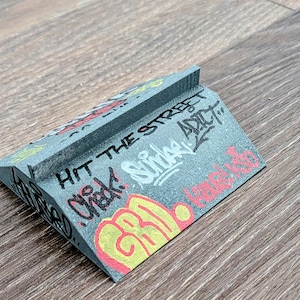 May include: A gray, miniature ramp with graffiti art. The ramp features the words "Hit the Street" and other graffiti tags in red, yellow, and white. The ramp is designed for fingerboarding or other small-scale activities.