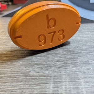May include: An orange, oval-shaped object with the letter "b" and the number "973" embossed on its surface. The object has a smooth texture and is set against a wooden background.