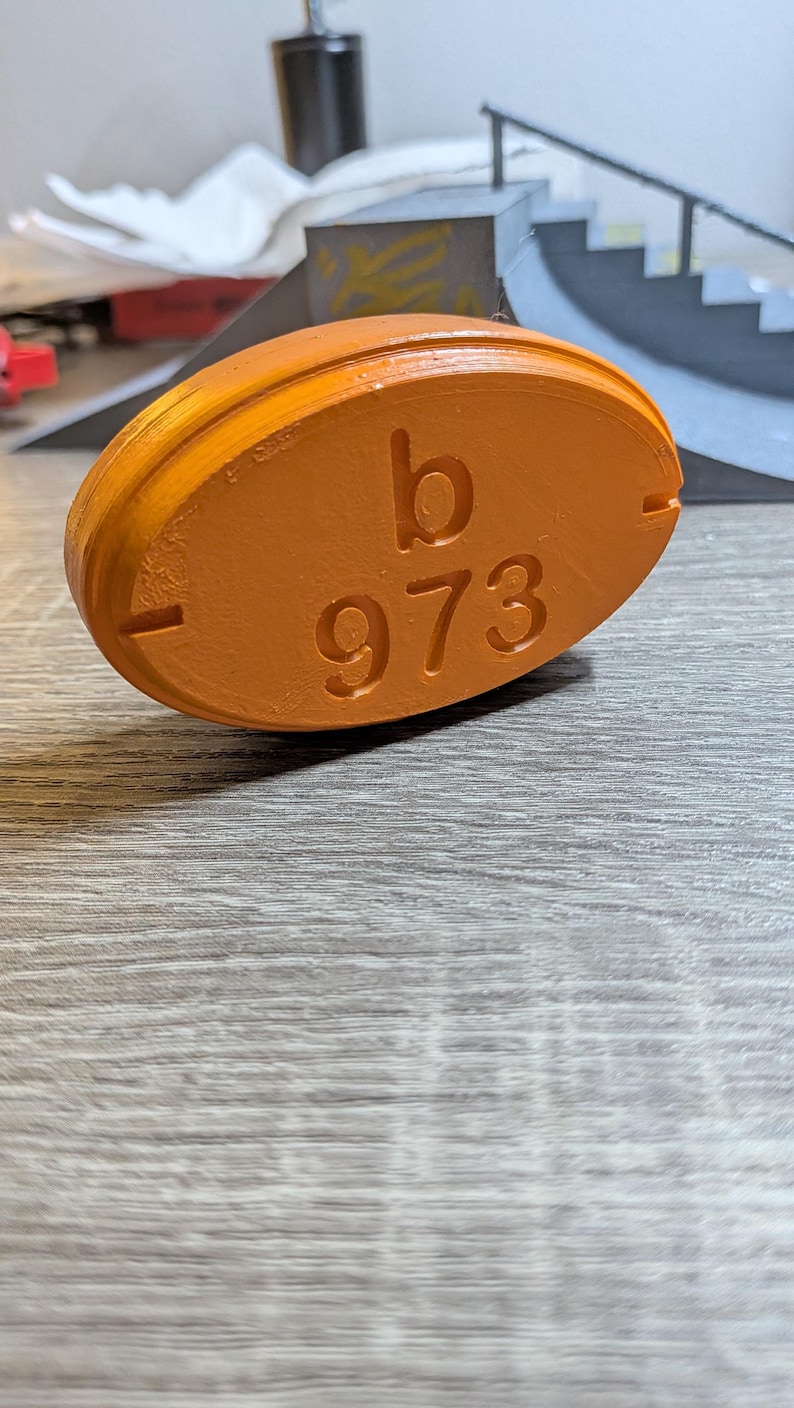 May include: An orange, oval-shaped object with the letter "b" and the number "973" embossed on its surface. The object has a smooth texture and is set against a wooden background.