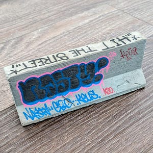 May include: A gray concrete barrier with graffiti art. The word "BEST" is in black and blue, outlined in pink. The text "HIT THE STREET" is above the word "BEST". Additional blue graffiti is below. The barrier is on a wood surface.