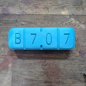 May include: A bright turquoise rectangular object with the letters "B 7 0 7" embossed on the top. The object is divided into four sections. The background is a wooden surface with various colors.