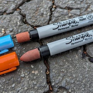 May include: Two grey Sharpie permanent markers with black text, one with the cap off, revealing a brown tip. Two caps, one blue and one orange, are next to the markers. The markers are on a grey, textured surface.