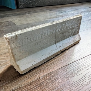 May include: A weathered, rectangular concrete block with a textured, wood-grain appearance. The block is primarily gray with a white stripe along the bottom edge. The object is likely a decorative or functional element for a garden or outdoor space.