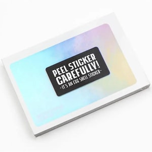 May include: A stack of white stickers with a holographic background. A black rectangular sticker with white text that reads "PEEL STICKER CAREFULLY! -IT'S AN EGG SHELL STICKER-" is affixed to the top of the stack.