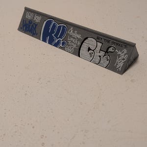 Puede incluir: Un rail de monopatín triangular gris con arte de graffiti. El rail presenta letras azules y blancas, incluyendo las palabras "Lunch Time" y "Hit the Street". El diseño tiene una estética de arte callejero.