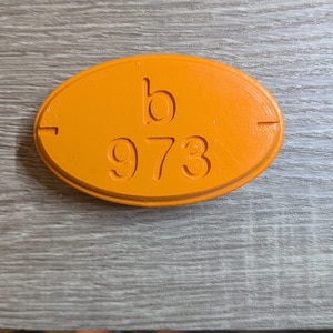 May include: An orange, oval-shaped tag with the letter "b" and the number "973" embossed on its surface. The tag has a slightly raised border and is set against a wooden background.