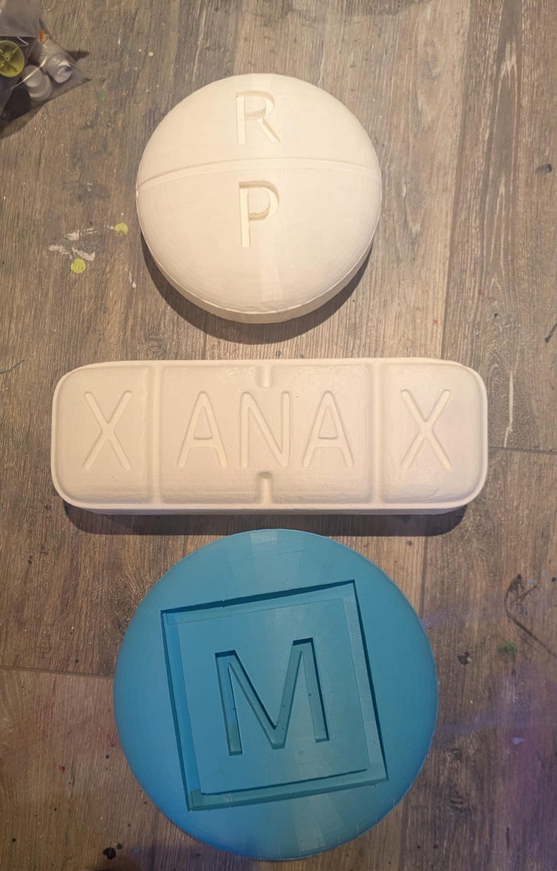 May include: Three white pill-shaped objects with raised lettering. The first object says "R P" on it. The second object says "XANAX" on it. The third object is round and says "M" on it. The third object is blue.