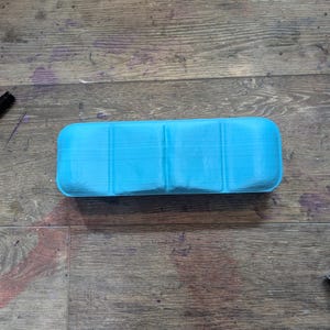 May include: A light blue plastic storage case with four compartments. The case is empty and has a smooth, slightly textured surface.