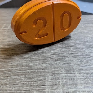 May include: An orange, oval-shaped object with the number "20" carved into its surface. The object is made of a solid material and has a smooth, matte finish. The background is a wooden surface.