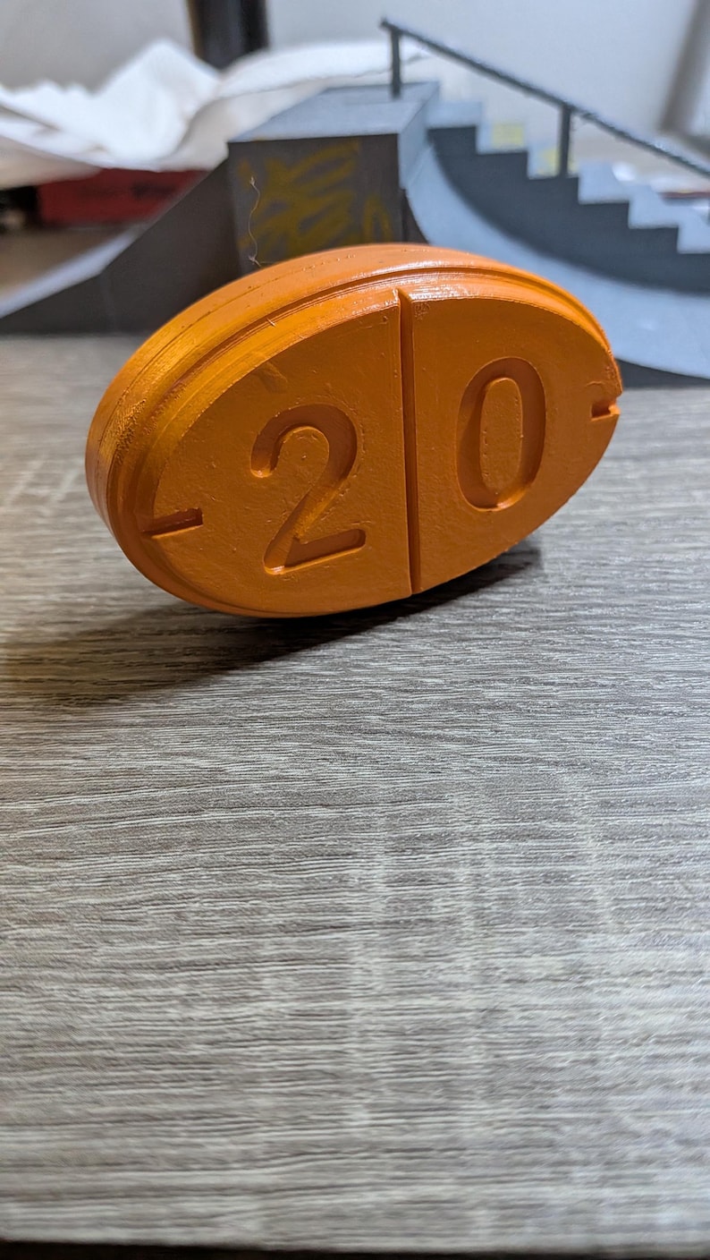 May include: An orange, oval-shaped object with the number "20" carved into its surface. The object is made of a solid material and has a smooth, matte finish. The background is a wooden surface.