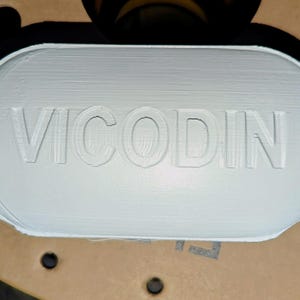 May include: White oval-shaped pill bottle with the word "VICODIN" embossed on the front.