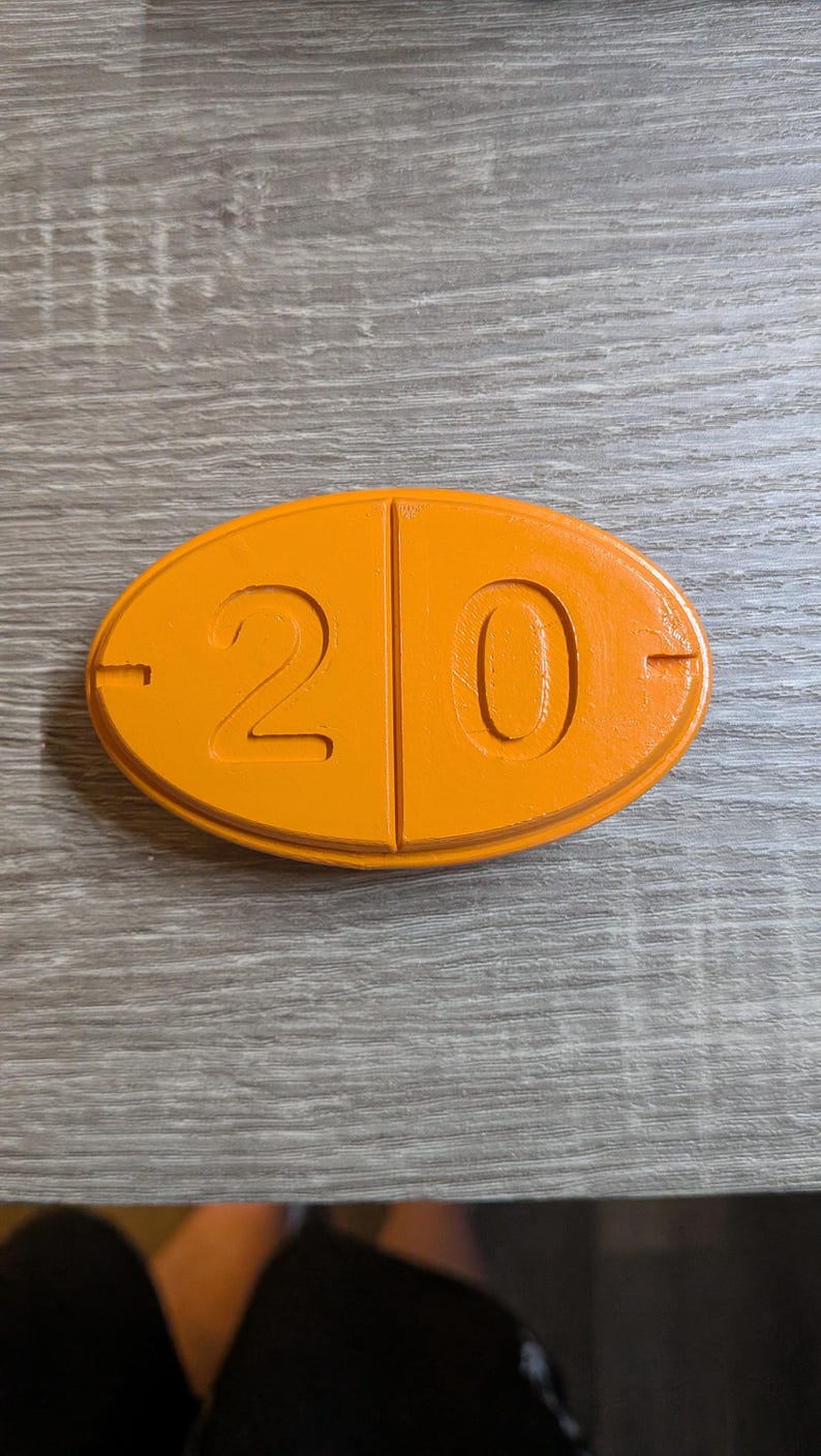 May include: An orange, oval-shaped object with the number "20" prominently displayed. The number is divided into two sections, with a horizontal line on each side. The object is on a wooden surface.