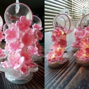 Peut inclure: Quatre bougeoirs en verre transparent avec des accents de fleurs de cerisier rose. Les bougeoirs sont allumés et les fleurs sont illuminées.