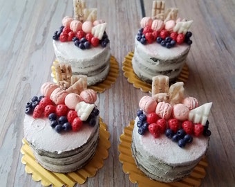 Peut inclure: Quatre gâteaux miniatures décorés de macarons roses et blancs, de baies rouges et bleues et de chocolat blanc. Les gâteaux sont sur des napperons en papier doré.