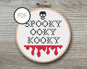 May include: A white embroidery hoop with a cross-stitch design that reads "SPOOKY OOKY KOOKY" with a skull above the text and red dripping blood below. The hoop is on a woodgrain background.