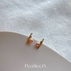 May include: A pair of gold earrings with a small pink pearl and a line of clear cubic zirconia stones. The earrings are on a white surface with the text "PearlfectX 2021 collection".