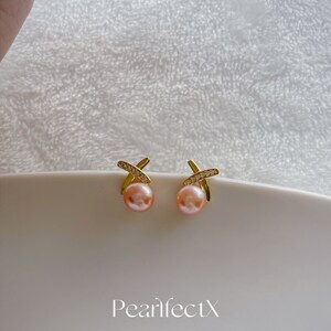 May include: A pair of gold earrings with a pearl and a cross design. The earrings are set with small clear stones. The earrings are on a white surface. The text "PearlfectX 2021 collection" is below the earrings.