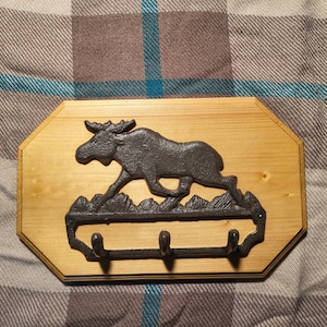 May include: A wooden wall-mounted coat rack with a moose silhouette design. The rack has three hooks and is made of natural wood.