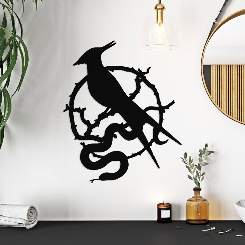A bird and snake inside a circle in black coloring are set against a white wall. A black surface with a candle and leaves inside a brown jar sits below the wall art. A gold lamp hangs above the wall art. A folded white towel sits off to the lower left corner.
