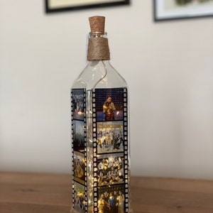 K&ouml;nnte beinhalten: Eine klare Glasflasche mit einem Korkverschluss und einer Schnur, die um den Hals gewickelt ist. Die Flasche ist mit Lichterketten und Fotos gef&uuml;llt, die auf Filmstreifen gedruckt sind.