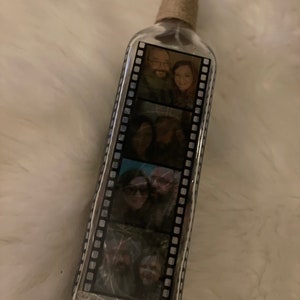 K&ouml;nnte beinhalten: Eine klare Glasflasche mit einem Korkverschluss und einer braunen Kordelwicklung. Die Flasche hat ein Filmstreifen-Design mit mehreren Fotos eines Paares im Inneren.