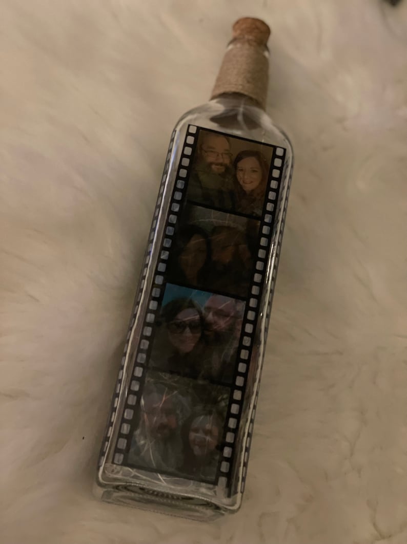 K&ouml;nnte beinhalten: Eine klare Glasflasche mit einem Korkverschluss und einer braunen Kordelwicklung. Die Flasche hat ein Filmstreifen-Design mit mehreren Fotos eines Paares im Inneren.