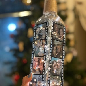 K&ouml;nnte beinhalten: Eine klare Glasflasche mit einem Korkverschluss und einer Schnur, die um den Hals gewickelt ist. Die Flasche ist mit schwarz-wei&szlig;en Filmstreifen mit Fotos von Personen darin verziert. Die Flasche ist mit wei&szlig;en Lichterketten beleuchtet.