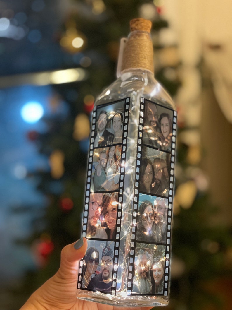 K&ouml;nnte beinhalten: Eine klare Glasflasche mit einem Korkverschluss und einer Schnur, die um den Hals gewickelt ist. Die Flasche ist mit schwarz-wei&szlig;en Filmstreifen mit Fotos von Personen darin verziert. Die Flasche ist mit wei&szlig;en Lichterketten beleuchtet.