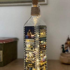K&ouml;nnte beinhalten: Eine klare Glasflasche mit einem Korkverschluss und einer Schnur, die um den Hals gewickelt ist. Die Flasche ist mit Fotos von Menschen in einem Filmstreifen-Design verziert. Die Flasche wird von innen mit wei&szlig;en Lichterketten beleuchtet.