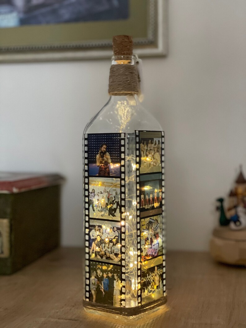 K&ouml;nnte beinhalten: Eine klare Glasflasche mit einem Korkverschluss und einer Schnur, die um den Hals gewickelt ist. Die Flasche ist mit Fotos von Menschen in einem Filmstreifen-Design verziert. Die Flasche wird von innen mit wei&szlig;en Lichterketten beleuchtet.