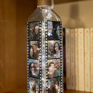 K&ouml;nnte beinhalten: Eine klare Glasflasche mit einer Schnur um den Hals. Die Flasche ist mit Lichterketten gef&uuml;llt und hat Fotos von Menschen auf der Au&szlig;enseite. Die Fotos sind in einem Filmstreifenmuster angeordnet.