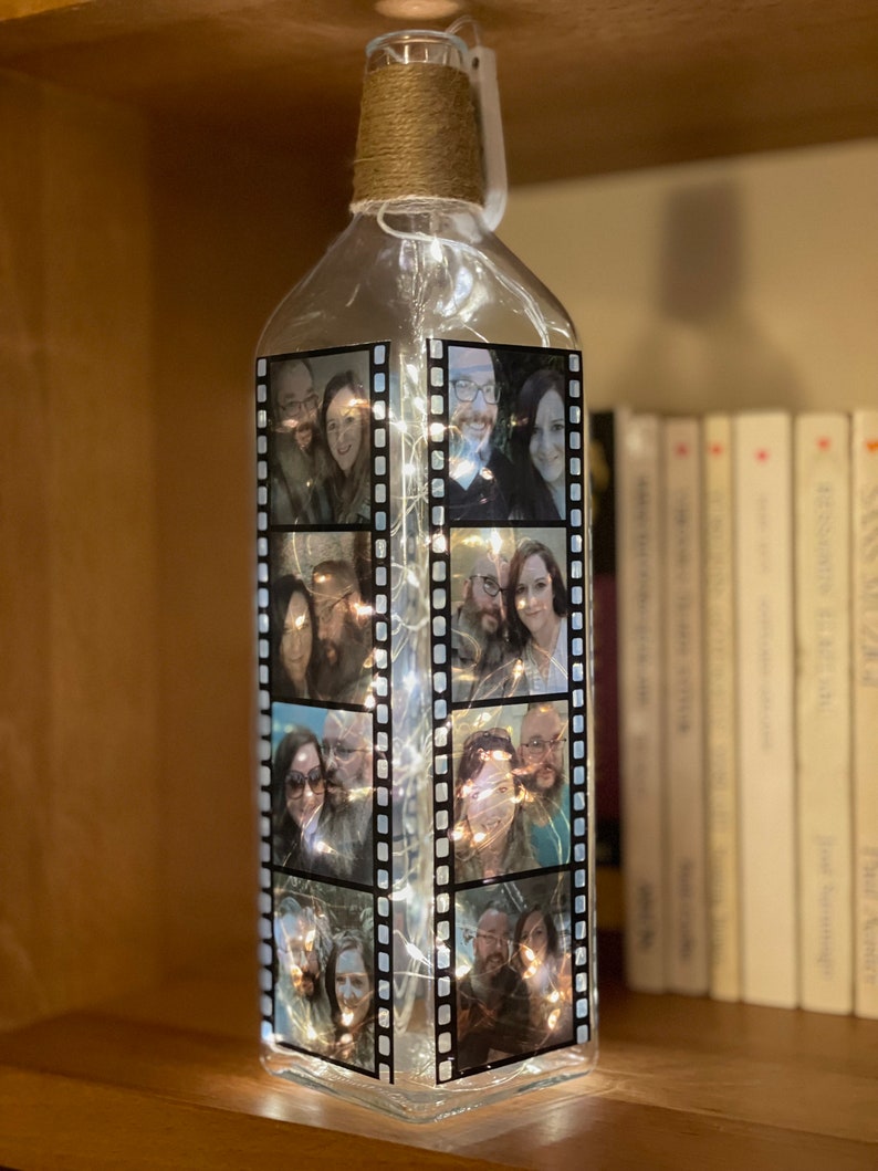 K&ouml;nnte beinhalten: Eine klare Glasflasche mit einer Schnur um den Hals. Die Flasche ist mit Lichterketten gef&uuml;llt und hat Fotos von Menschen auf der Au&szlig;enseite. Die Fotos sind in einem Filmstreifenmuster angeordnet.