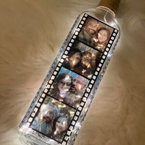K&ouml;nnte beinhalten: Eine klare Glasflasche mit einem Korkverschluss und einer Lichterkette im Inneren. Die Flasche ist mit einem Schwarzwei&szlig;-Filmstreifenmuster und Fotos eines Paares verziert.
