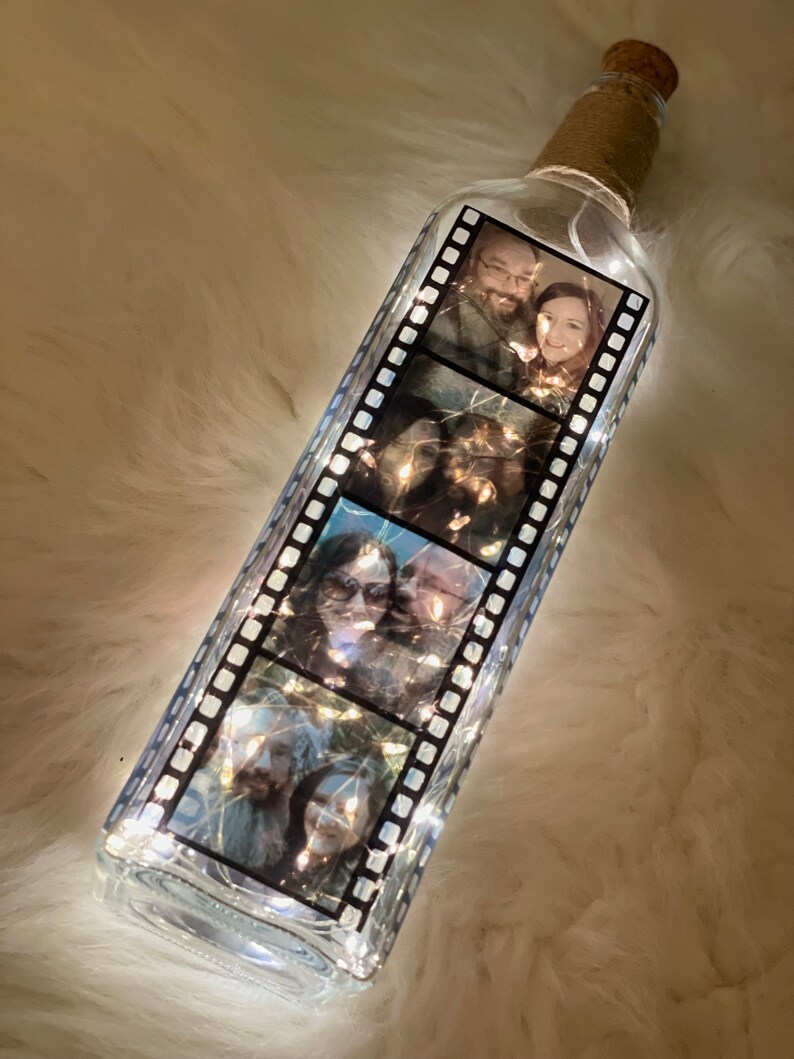 K&ouml;nnte beinhalten: Eine klare Glasflasche mit einem Korkverschluss und einer Lichterkette im Inneren. Die Flasche ist mit einem Schwarzwei&szlig;-Filmstreifenmuster und Fotos eines Paares verziert.