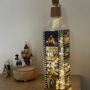 K&ouml;nnte beinhalten: Eine klare Glasflasche mit einem Korkverschluss und einer Schnur, die um den Hals gewickelt ist. Die Flasche ist mit Lichterketten gef&uuml;llt und hat Fotos von Menschen, die auf der Au&szlig;enseite gedruckt sind. Die Fotos sind in einem vertikalen Streifen angeordnet, der an einen Filmstreifen erinnert.
