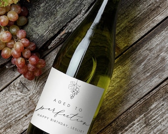 May include: A bottle of white wine with a custom label that reads "Aged to Perfection" and "Happy Birthday, Leslie!" in elegant script. The label features a simple grape illustration. Grapes and a leaf are next to the bottle on a rustic wooden surface.
