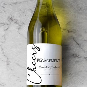 May include: A clear glass wine bottle featuring a white rectangular label with black text that reads Cheers TO YOUR ENGAGEMENT Hannah & Michael 3.22.2025 with a small heart icon. The bottle contains a pale yellow liquid and has a tall, slender neck.