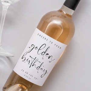 May include: A bottle of white wine with a black cap and a white label that reads "Cheers to your golden birthday" in black script. A wine glass is in the background.