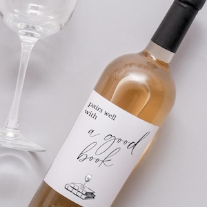 May include: A bottle of rosé wine with a black cap and a white label that reads "pairs well with a good book." A wine glass is in the background. The wine bottle is angled diagonally.