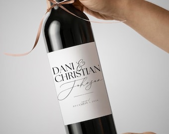 May include: A dark wine bottle with a white label featuring the names "DANI & CHRISTIAN" and the last name "Johansen" in elegant script. The label also includes the date "DECEMBER 1, 2024". A light brown ribbon is tied around the neck of the bottle.