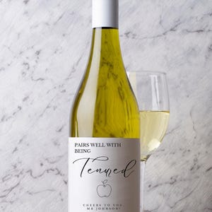 May include: A bottle of white wine with a white label that reads "Pairs well with being Tenured" with an apple graphic. A wine glass filled with wine is next to the bottle. The background is a marble pattern.