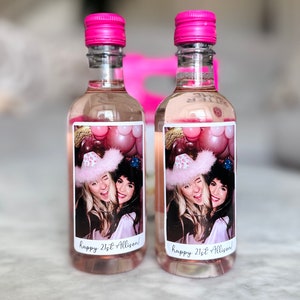May include: Two clear glass bottles with pink liquid and pink caps. Each bottle has a label with a photo of two women and the text "happy 21st Allison!" The background is blurred.