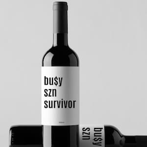 May include: Two black wine bottles with white labels. The labels read "busy szn survivor" in a bold, sans-serif font. One bottle is upright, and the other is lying down. The background is a plain, light gray.