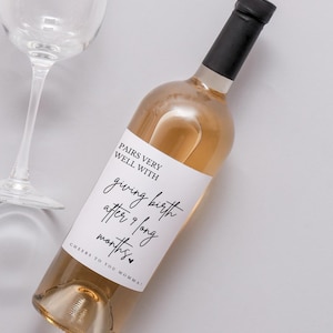 May include: A bottle of rosé wine with a black cap and a white label. The label reads "Pairs very well with giving birth after 9 long months. Cheers to you Momma!" A wine glass is in the background.