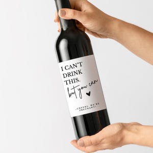May include: A dark glass wine bottle featuring a white rectangular label with black text that reads: I CAN'T DRINK THIS. but you can. A small black heart icon is included, followed by the text: -CHEERS, WE'RE PREGNANT!